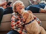 A happy family is sitting in the living room. Their son is sitting on the floor with the family dog licking his face.
